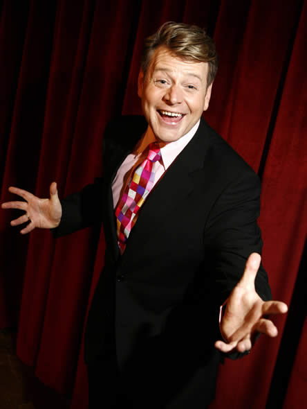 Mark Kristian in a black suit and a colourfull tie smiling from ear to ear, on a stage with a rich red backdrop